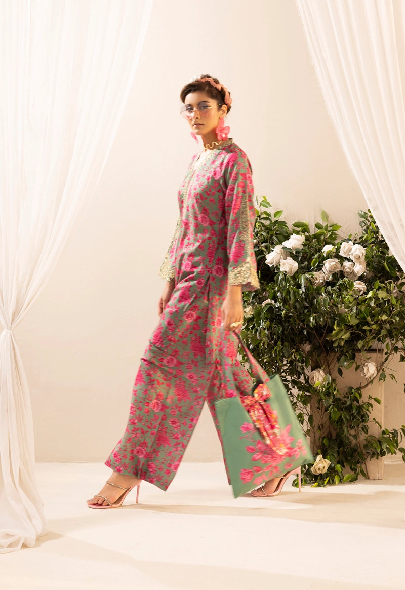 Woman in a floral outfit holding a green bag against a white curtain and plant backdrop
