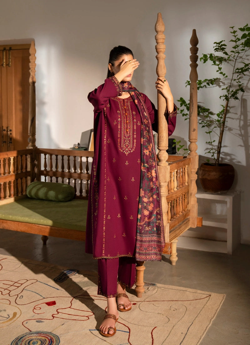 Woman in a traditional maroon outfit with floral patterns standing in a room with wooden furniture and plants.