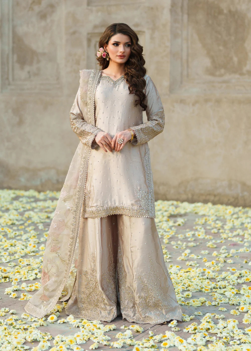 Woman in a traditional outfit standing on a ground with flowers, against a textured wall.